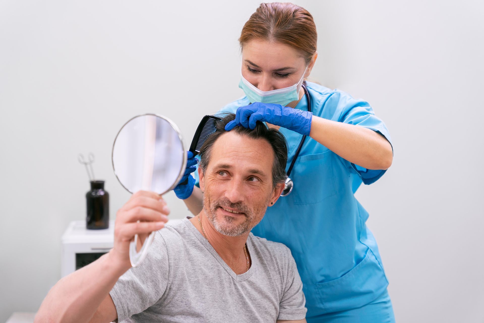 A medical professional combs a man's hair in a clinic while he holds a mirror, examining his hair.