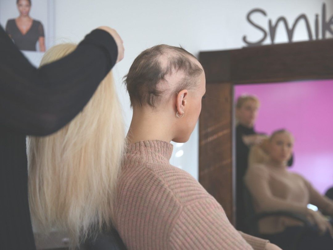 Woman with hair loss, holding a blonde wig in front of her head at a salon.