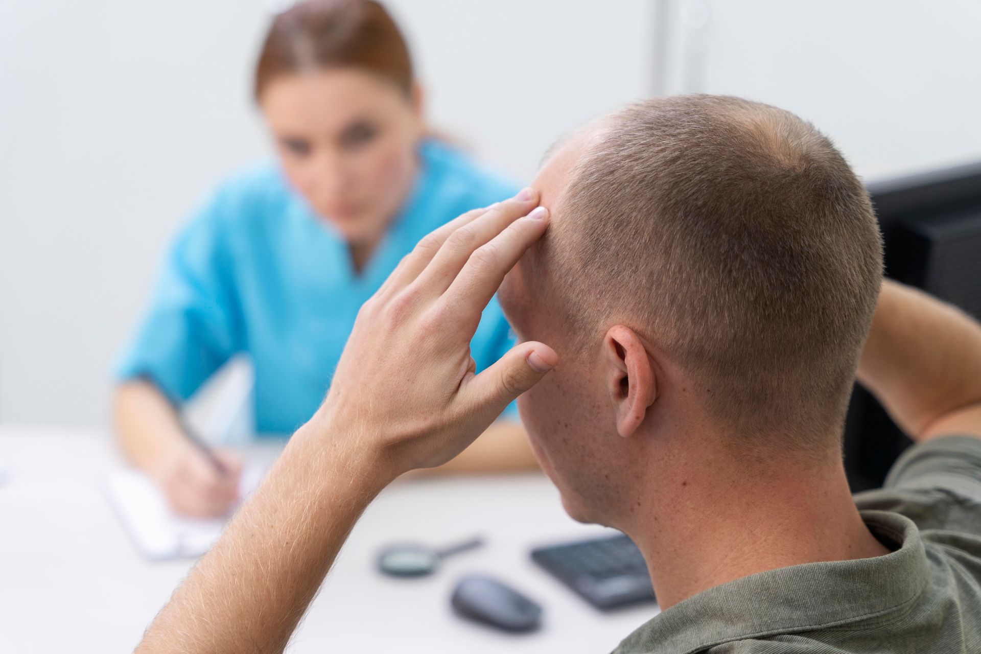Man at a desk holding head, talking to a person taking notes. White desk, blue shirt.