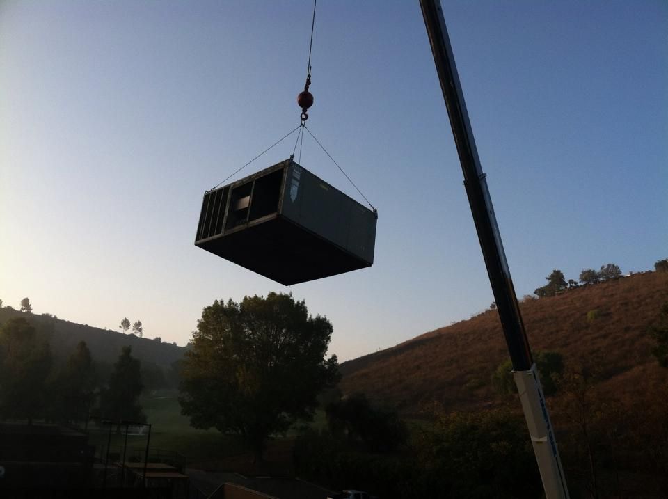 Crane lifting a large, rectangular HVAC unit outdoors at dusk. Hills and trees in the background.