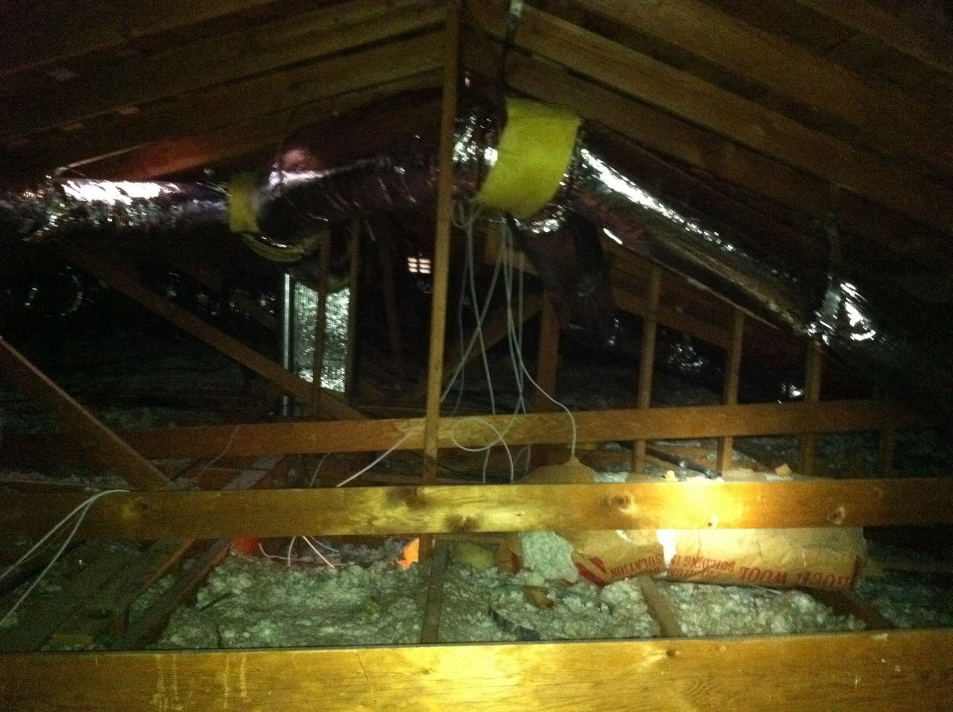 Attic with ductwork, insulation, and wooden beams. Dark, interior space.