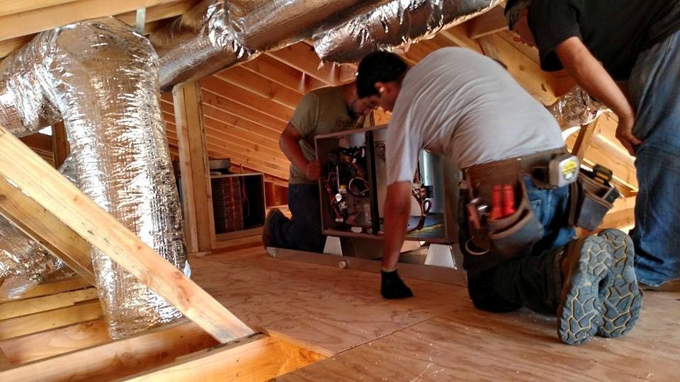 Three workers installing HVAC unit in an attic. Man in center, kneeling, working on the unit. Ductwork visible.
