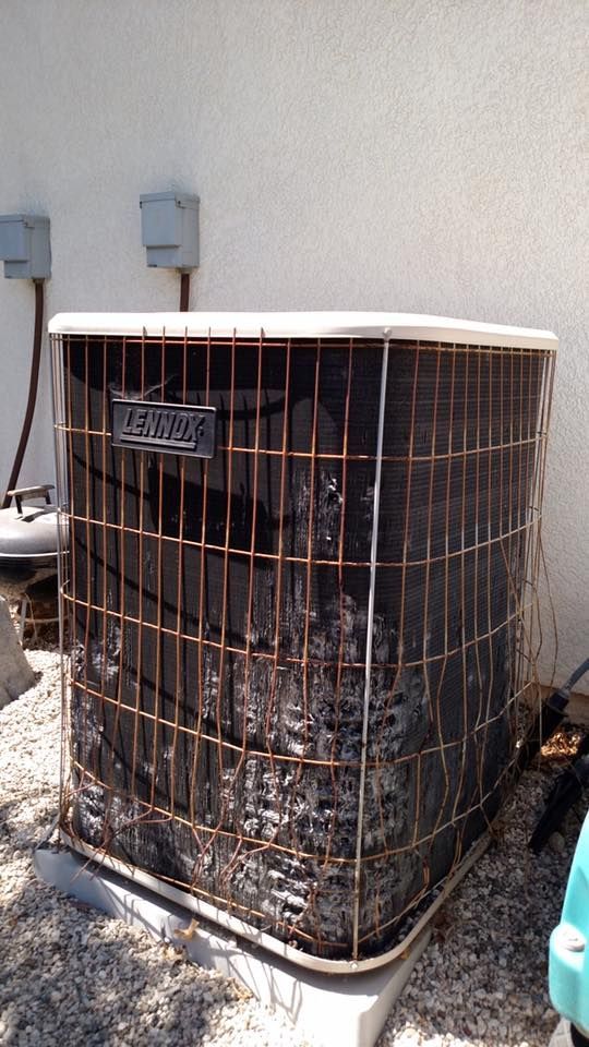 Outdoor air conditioning unit, dark metal, with surrounding metal grill, set against a white wall.
