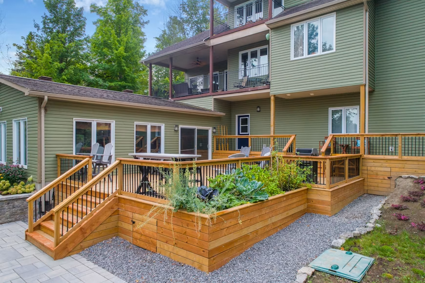 Home exterior with wooden deck, stairs, and raised garden beds beside a green house.