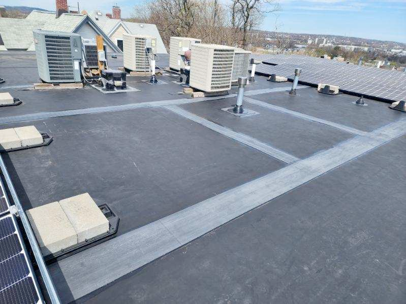 Black flat roof with HVAC units and solar panels. Grey pathways and tan brick weights are visible.