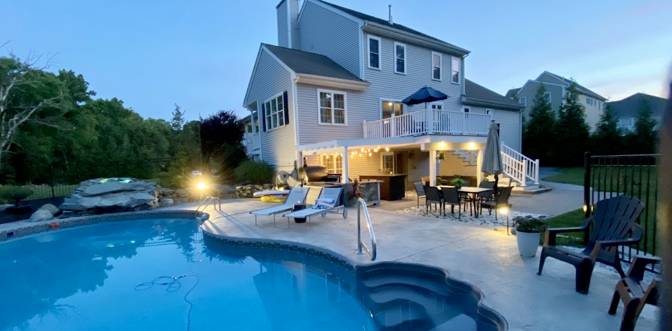 Backyard with pool, house, deck, chairs, and table lit by soft evening lights.