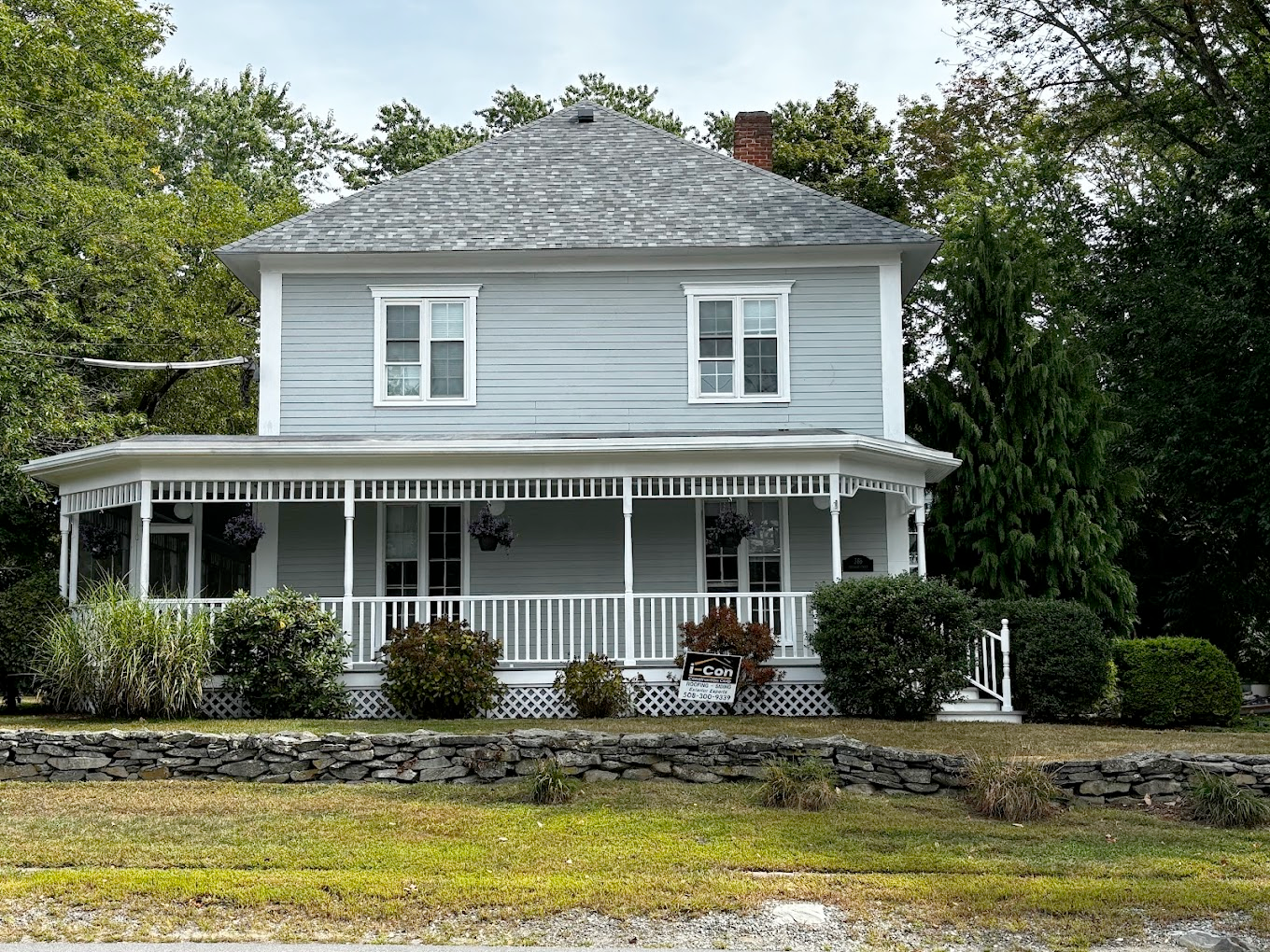 Two-story blue house with wrap-around porch, surrounded by greenery and a stone wall.