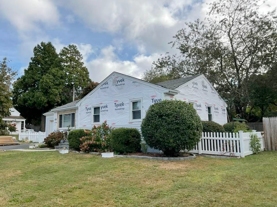 House exterior wrapped in white Tyvek, green lawn, white picket fence, trees and cloudy sky.