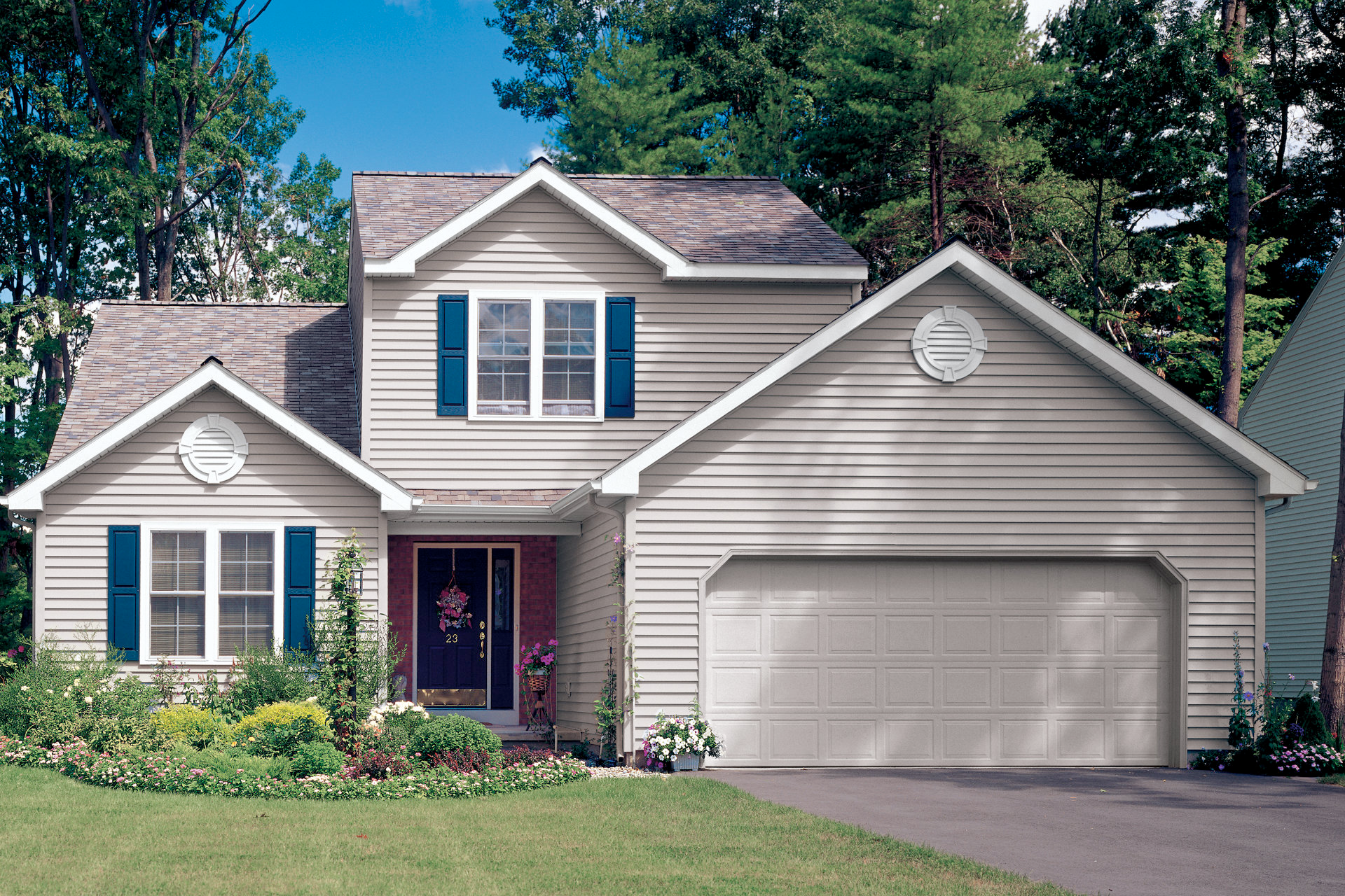 Beige two-story house with blue shutters and a two-car garage. Green lawn and trees surround.