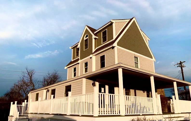 Three-story house with wrap-around porch, white railings, gray siding, and olive green roof against a cloudy sky.