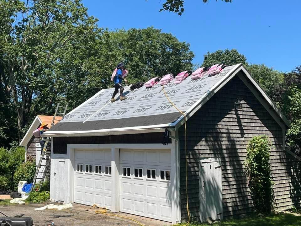 Roofers replacing a gray roof on a white garage with two doors, in a sunny yard.