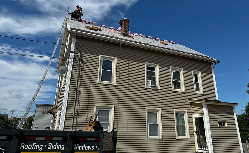 Roofers on a house replacing the roof on a sunny day.  Ladder, dumpster, and blue sky visible.
