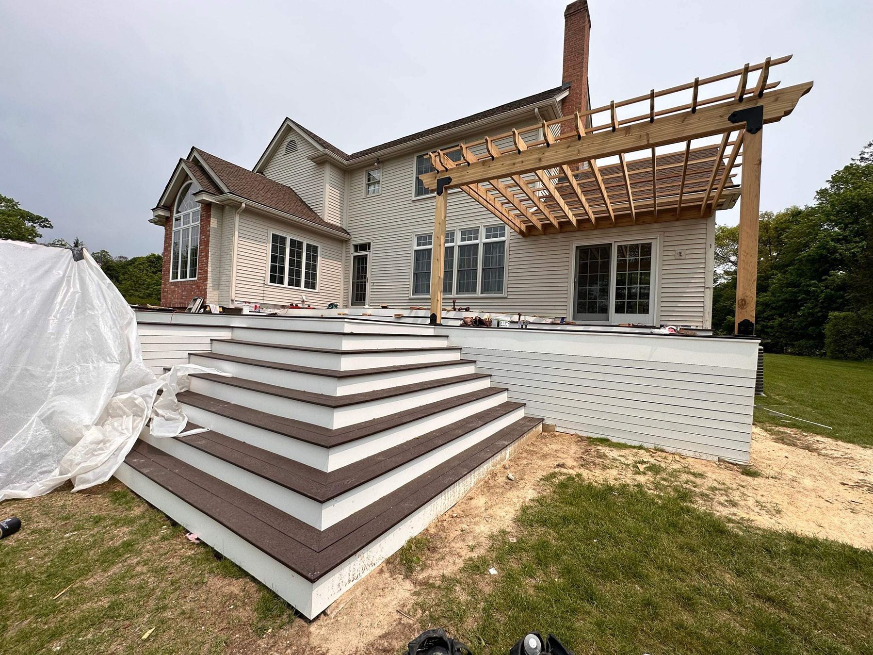 Back of a house with a deck under construction. White siding, brown stairs, pergola, and green grass.