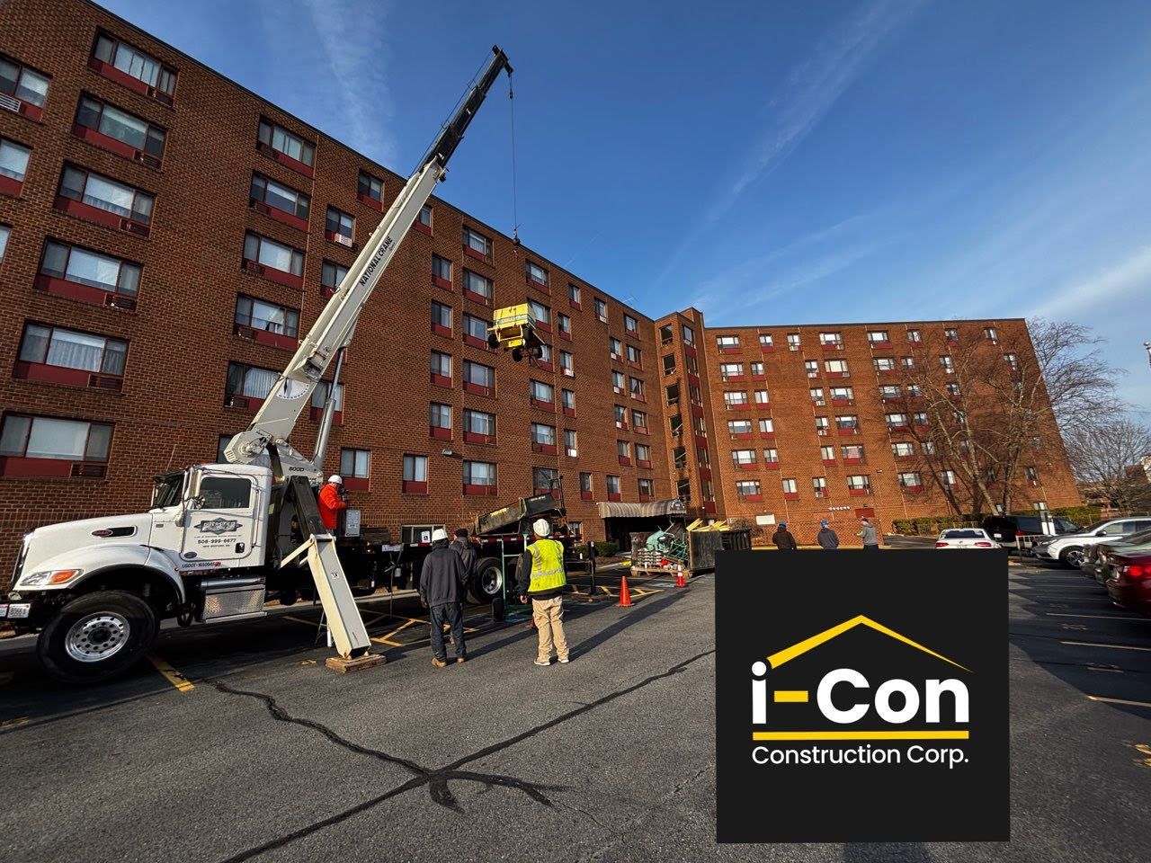 A crane lifts material at a multi-story brick building during construction. Construction workers are visible.