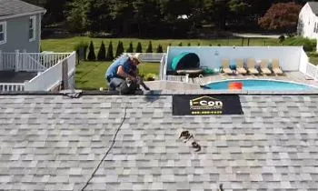 A worker kneeling on a shingled roof, installing roofing materials near an I-Con sign, overlooking a backyard with a pool.