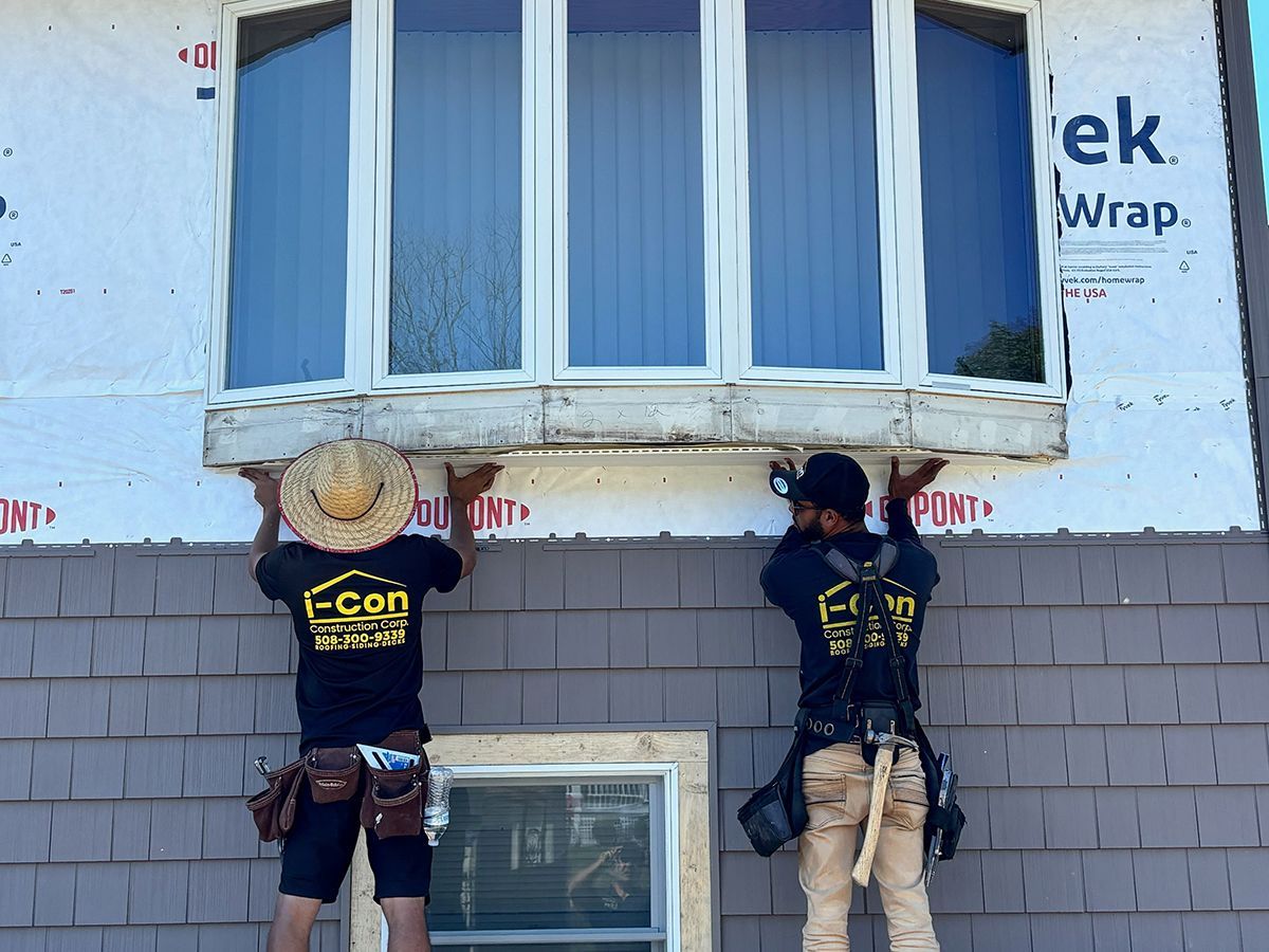 Two workers installing a window, wearing work attire, on a house with gray siding.