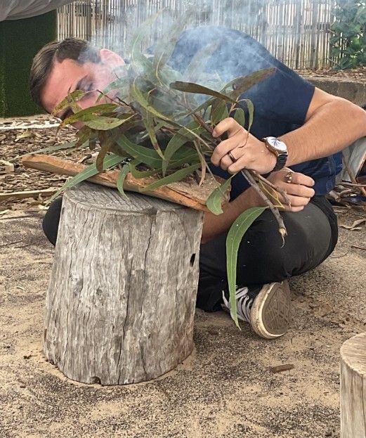 Man kneeling by a tree stump, blowing smoke from burning leaves, outdoors.