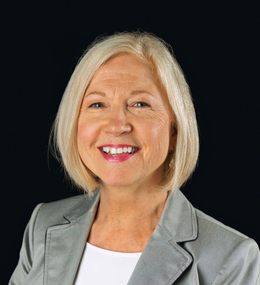 A woman with blonde hair is wearing a grey jacket and smiling.