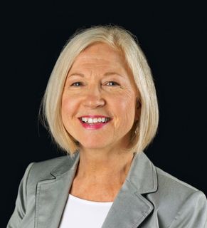 A woman with blonde hair is wearing a grey jacket and smiling.