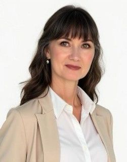 A woman in a tan jacket and white shirt is standing in front of a white background.