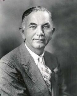 A man in a suit poses for a formal portrait; light skin, graying hair, and a patterned tie.