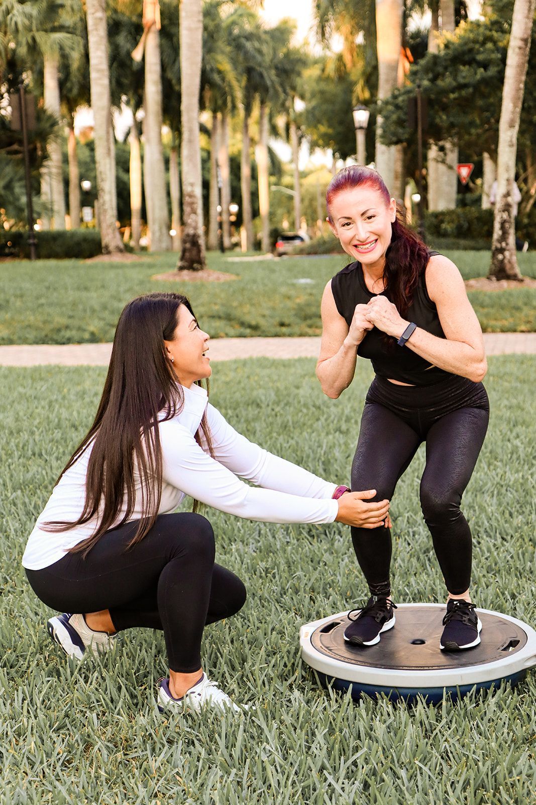 Two women are squatting on a balance board in a park.