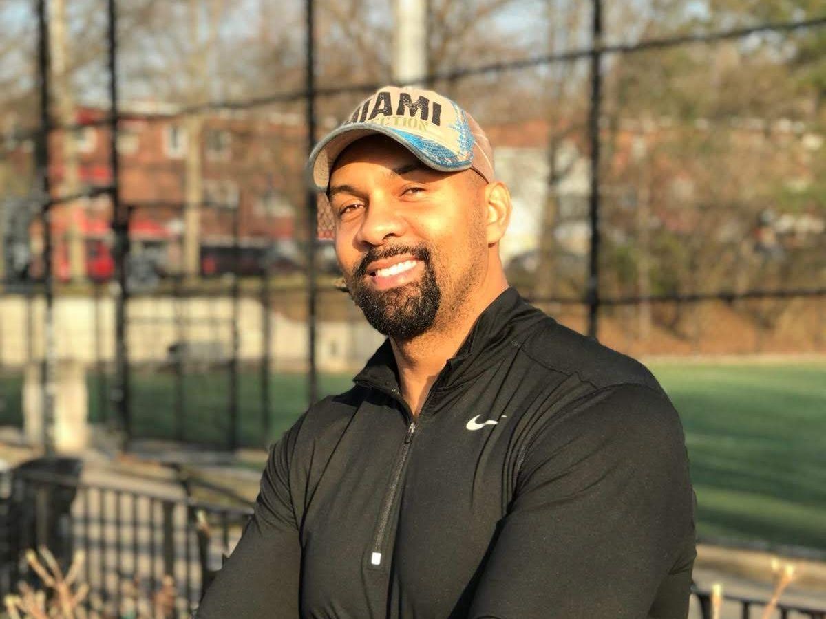 A man wearing a hat and a black shirt is standing in front of a fence.