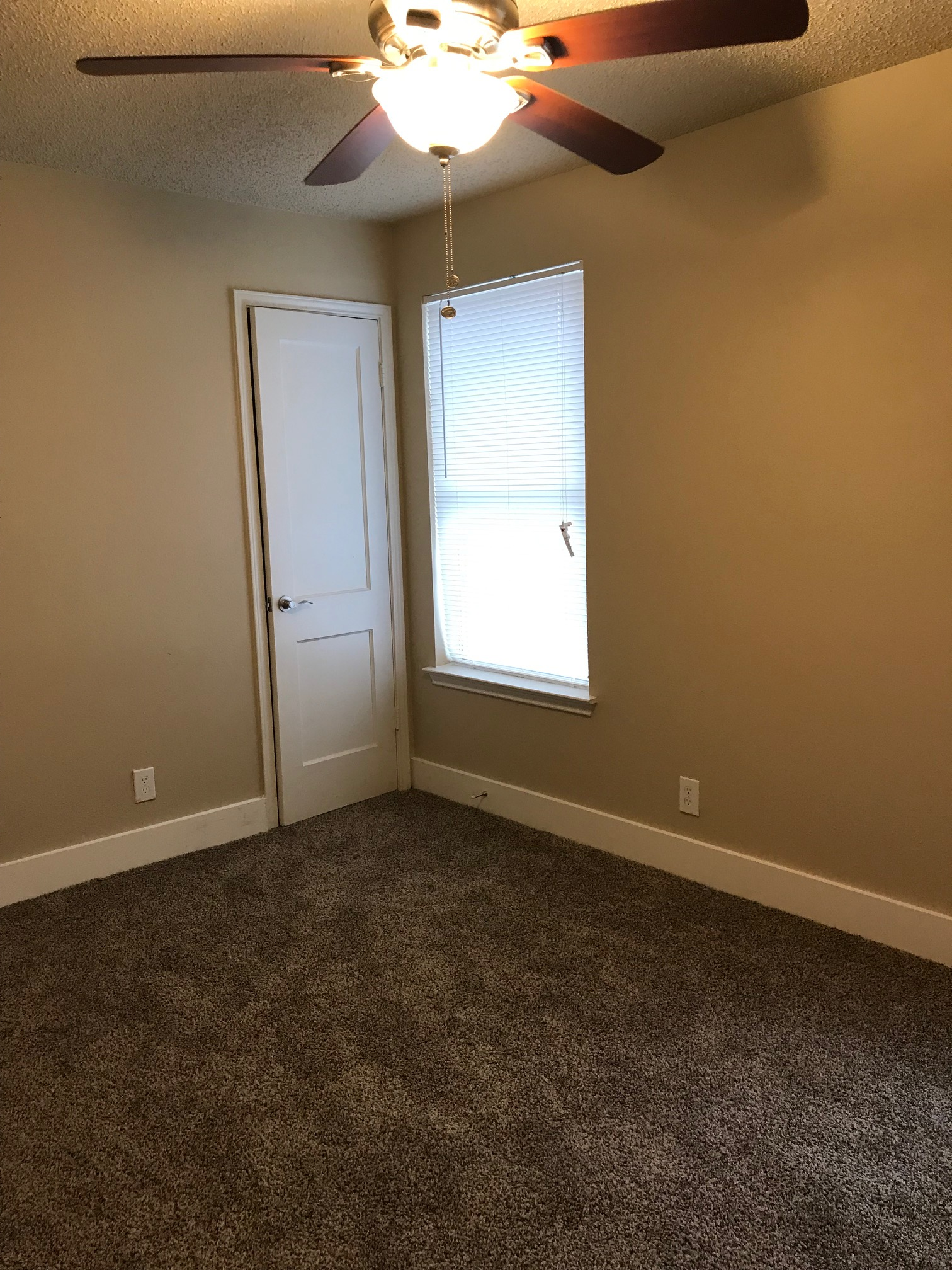 Empty bedroom with tan walls, brown carpet, white trim, door, and window. Ceiling fan with lights.