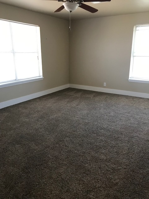 Empty room with brown carpet, beige walls, two windows, and a ceiling fan.