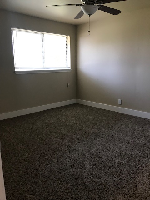 Empty room with beige walls, brown carpet, and a window. Ceiling fan is visible.