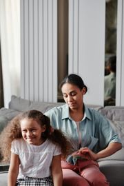 A woman is brushing a little girl 's hair while sitting on a couch.