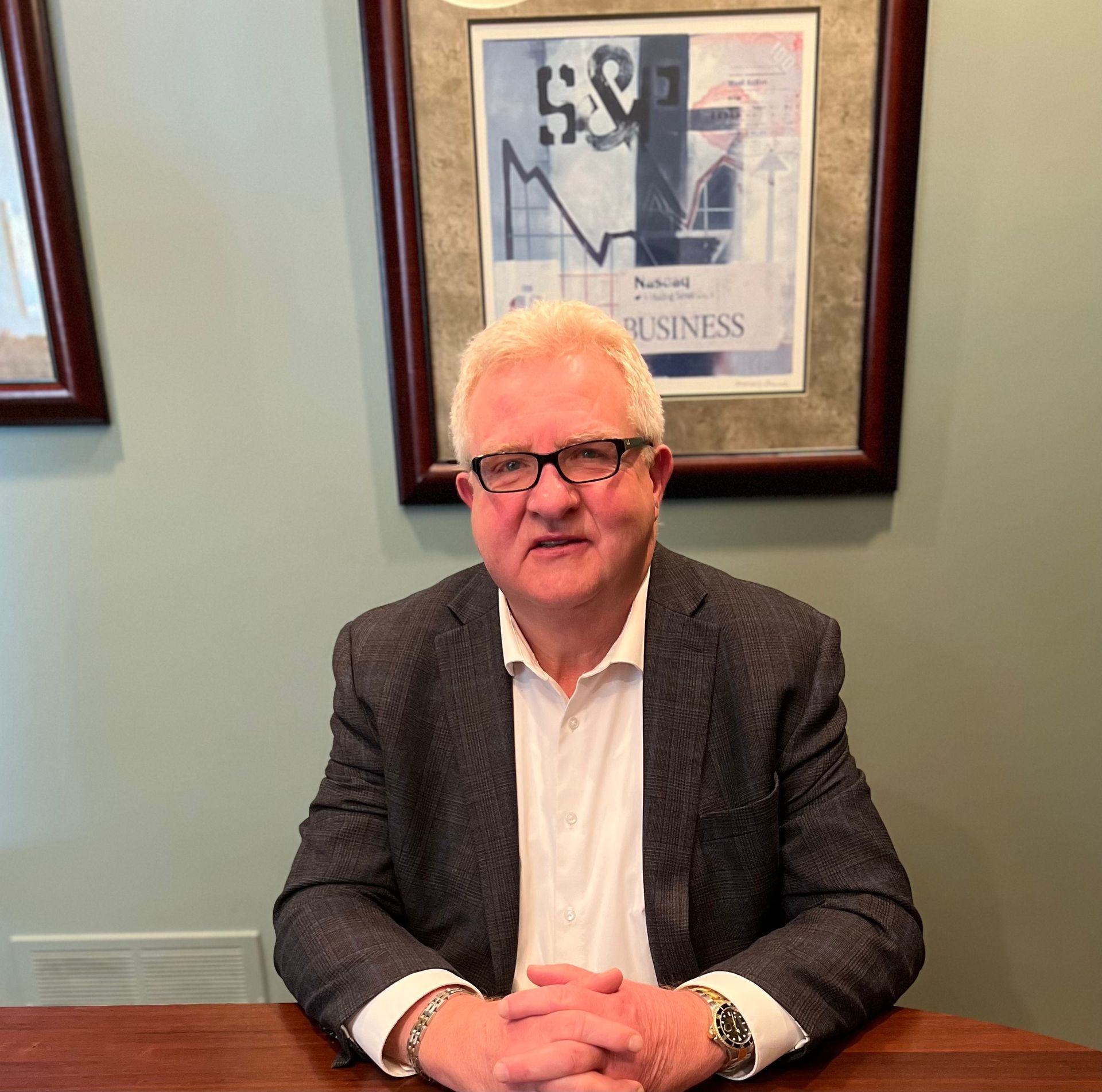 A man in a suit and glasses is sitting at a table with his hands folded
