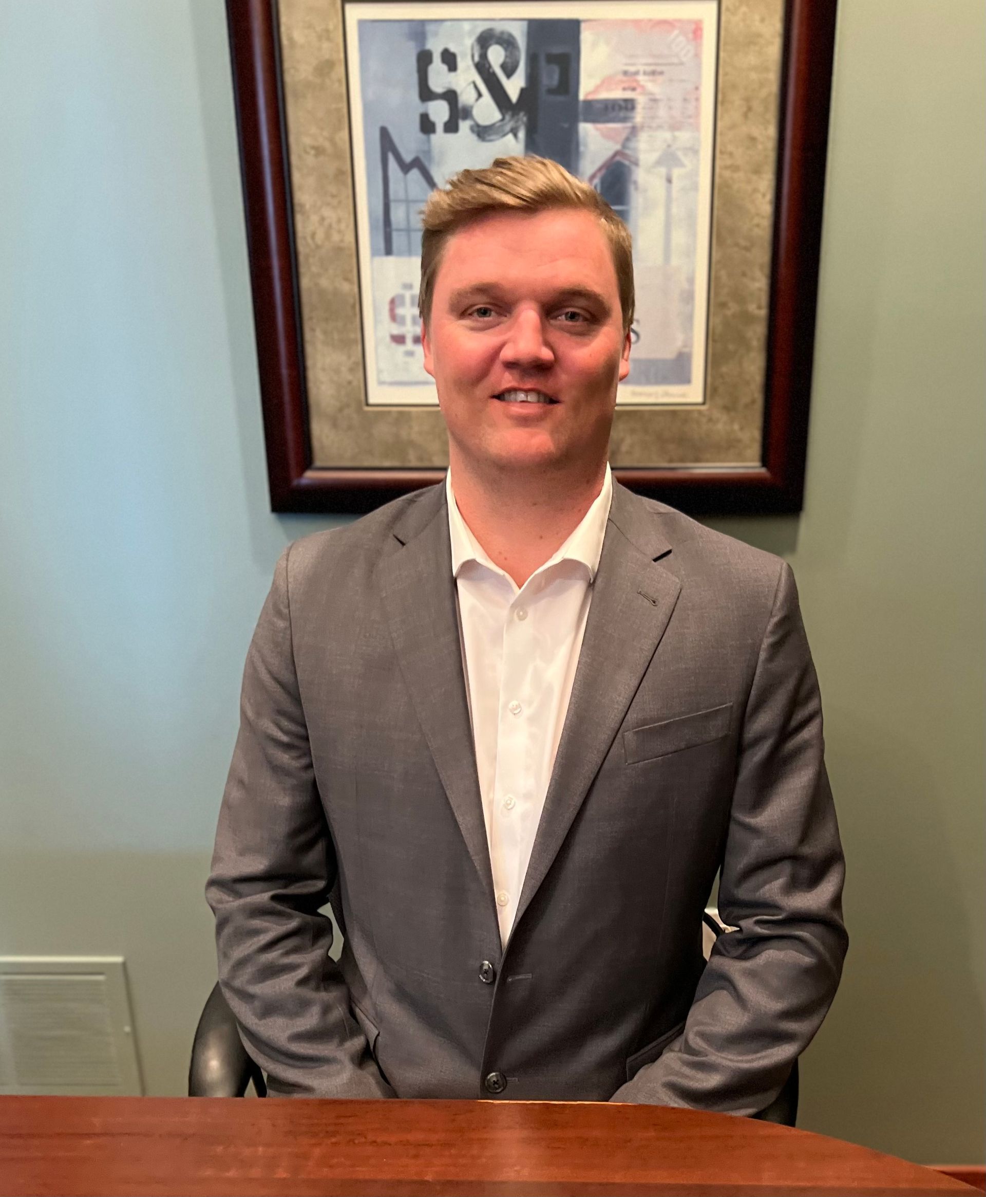 A man in a suit is sitting at a desk in front of a framed picture with the number 59 on it