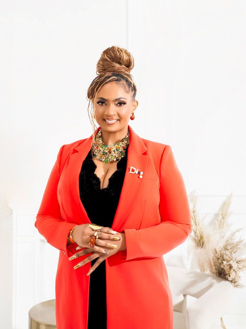 Woman in a red blazer, standing indoors, smiling. She wears a black top, jewelry, and has her hair up.