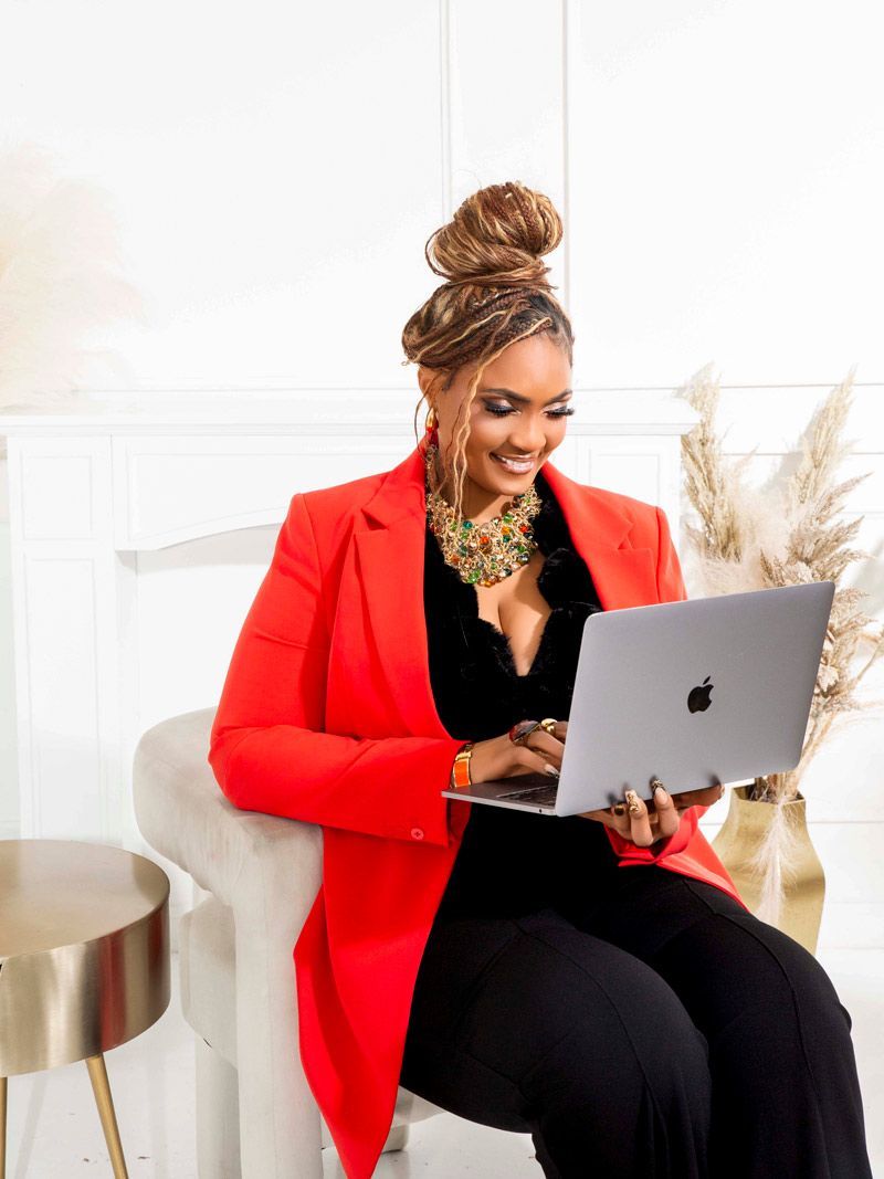 Woman in red blazer smiles while using a laptop, seated on a white chair.