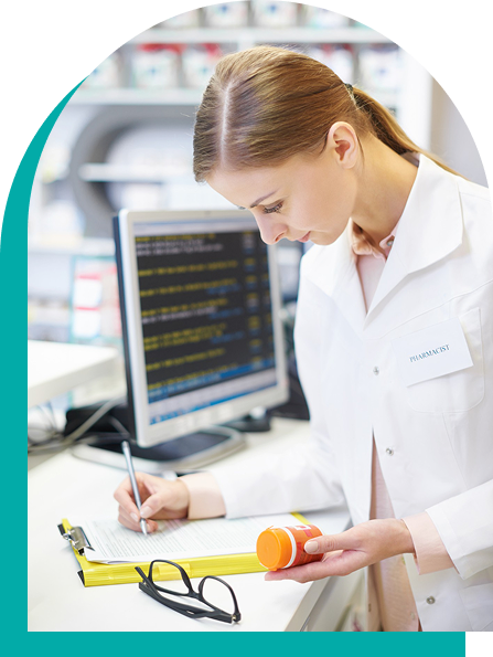 Pharmacist in lab coat reviewing prescription, holding pill bottle, with computer and glasses on counter.