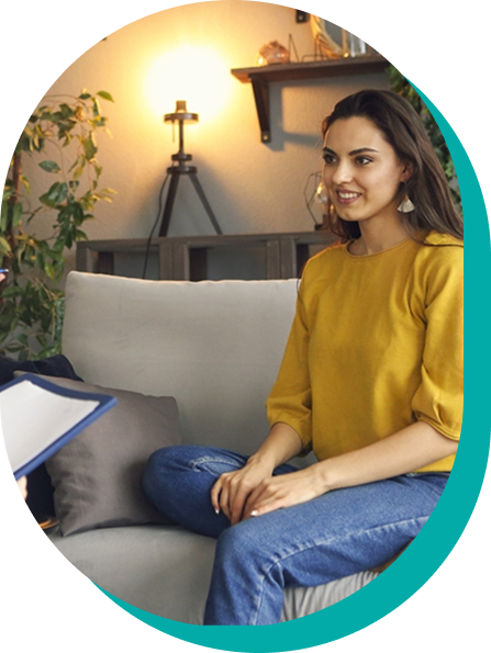 Woman in yellow top, blue jeans sitting on couch, smiling. Indoors, lit lamp in background.