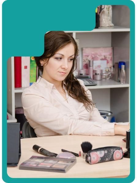 Woman at desk with makeup. Light-colored blouse, looking down, makeup products on desk, office setting.