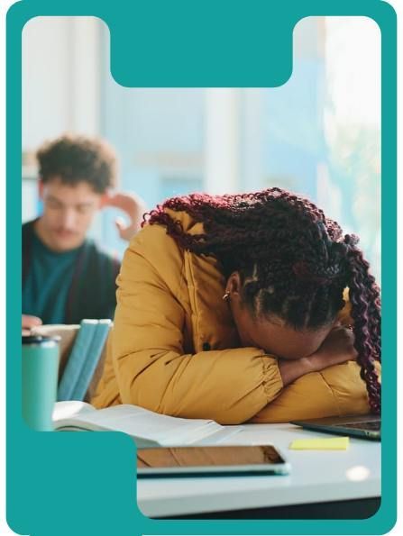 Woman slumped over a desk, face down, in a library setting. A student sits in the background.
