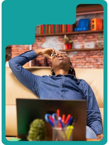 Person on couch looking up, hand on forehead, laptop in front. Bookshelf in background.