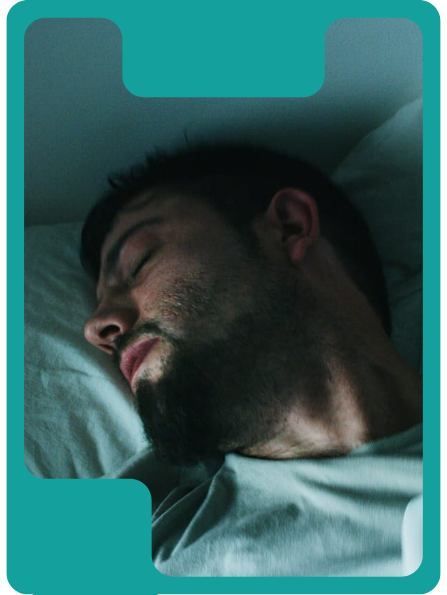 A man asleep in bed, side profile. He has a beard and is wearing a gray shirt.