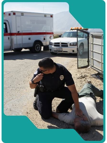 Police officer kneels over a person on the ground, likely injured. Ambulance and police car are in the background.