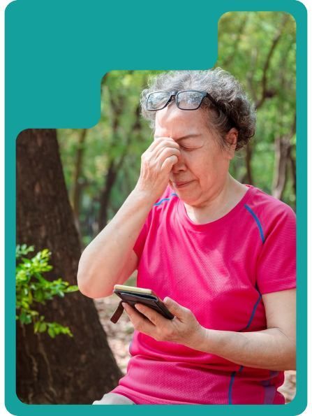 Woman in pink shirt holding phone, rubbing eye, outdoors.