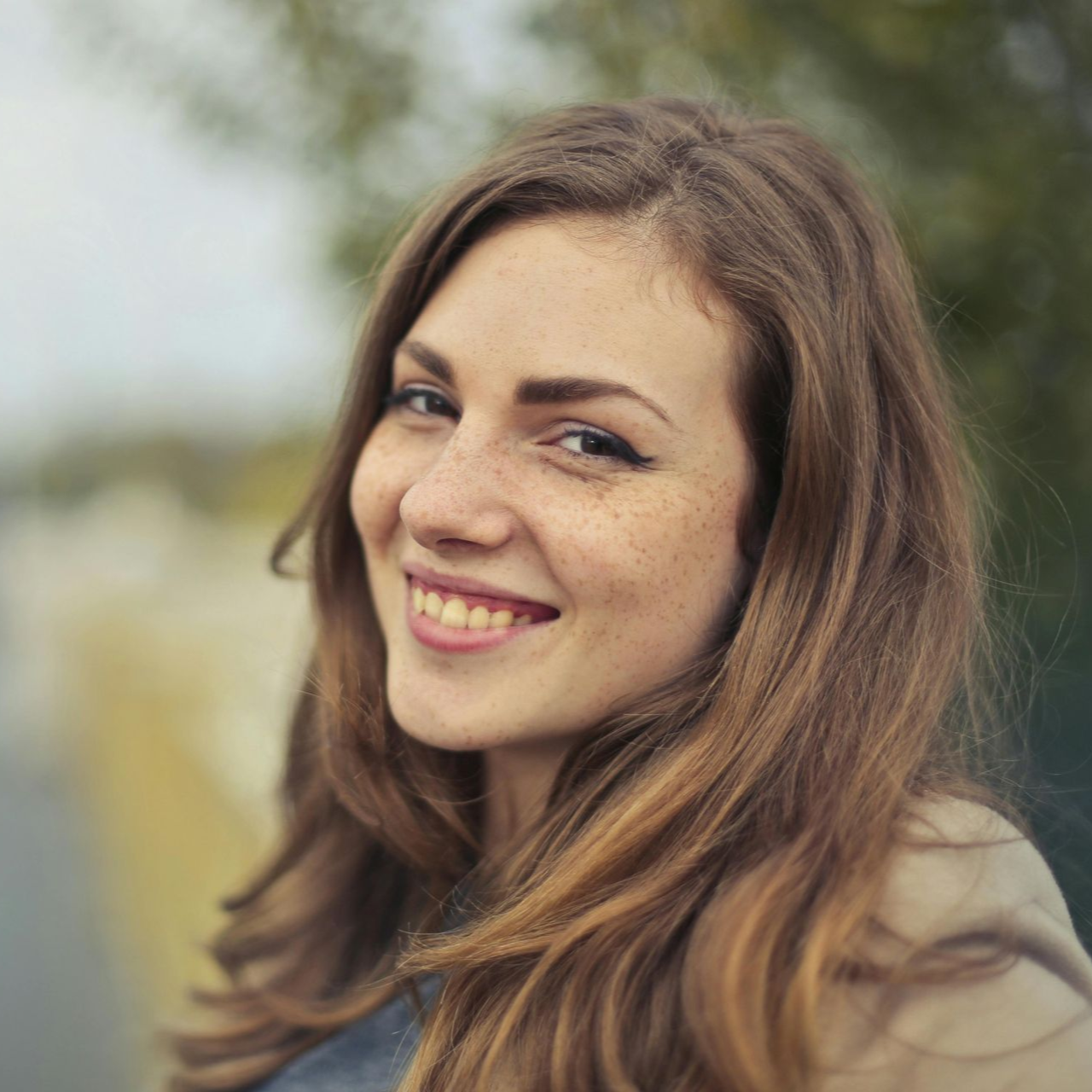 Smiling woman with long brown hair and freckles, outdoors with a blurred background.