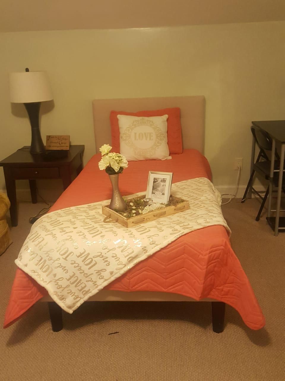 Coral-colored bed with throw and decorative items, lamp on nightstand. Beige walls and carpet.