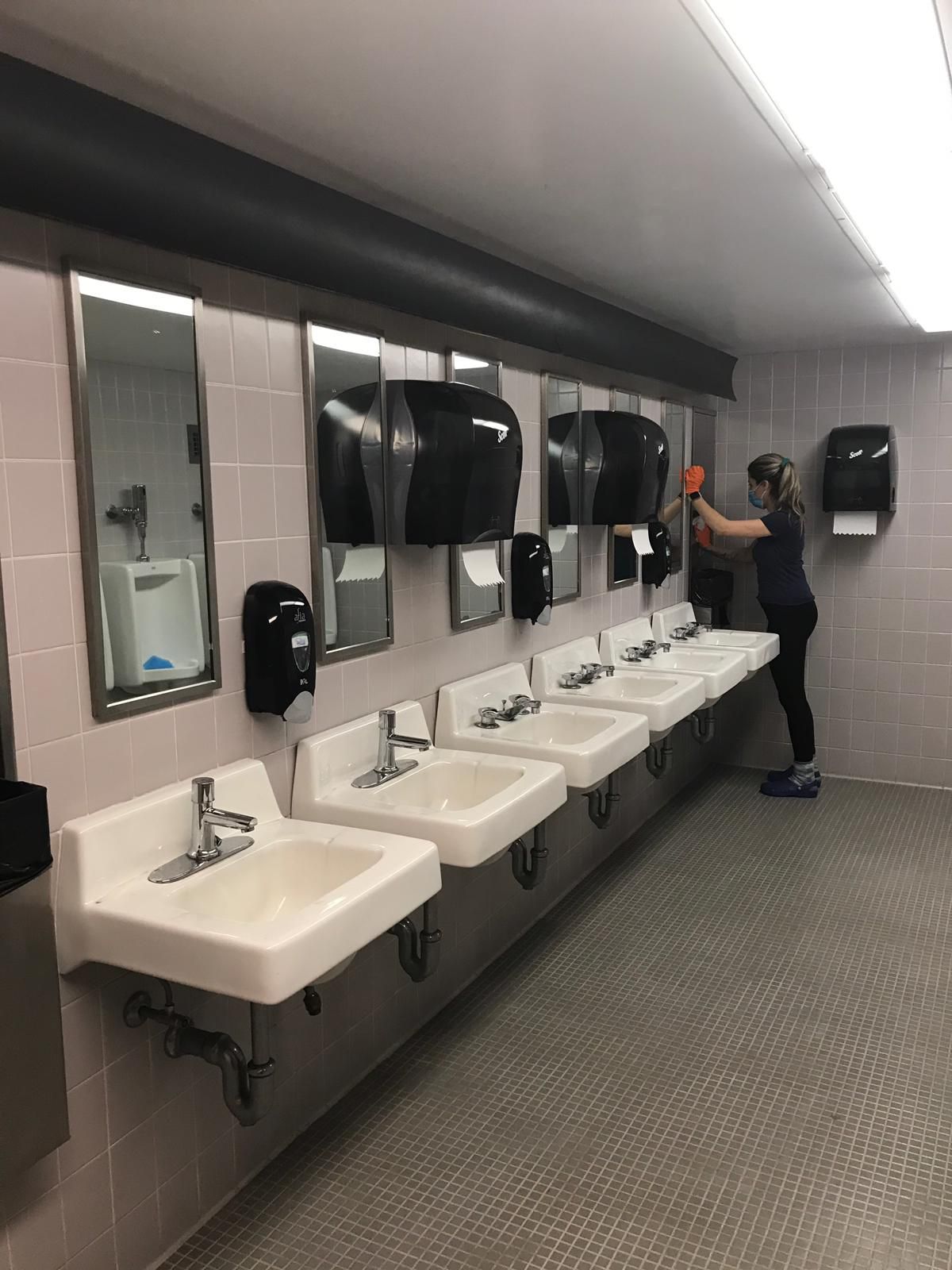 A person cleaning a row of bathroom sinks. White sinks and mirrors line the wall.