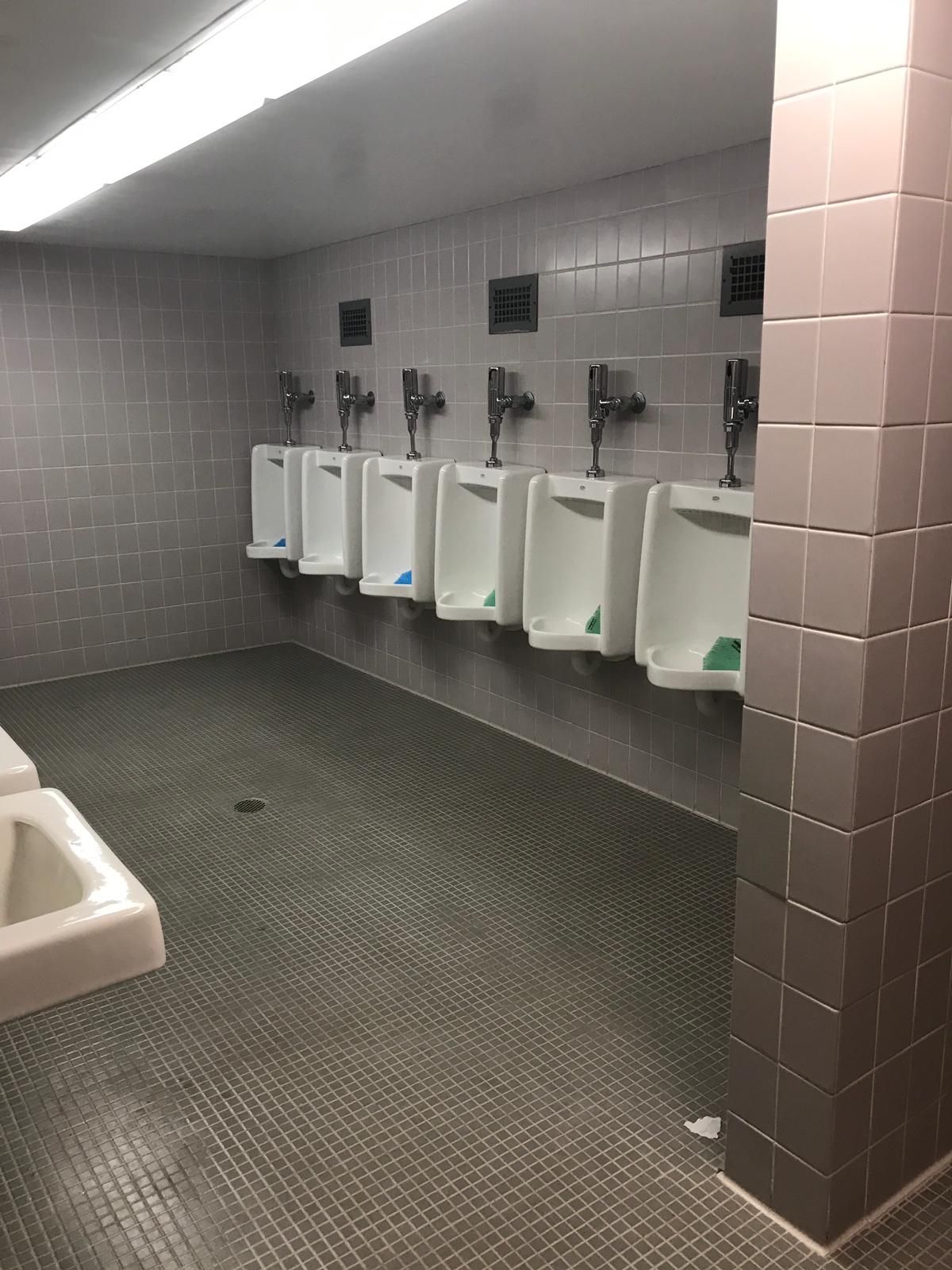 Public restroom with six urinals against gray tiled wall, floor is also gray tiles.