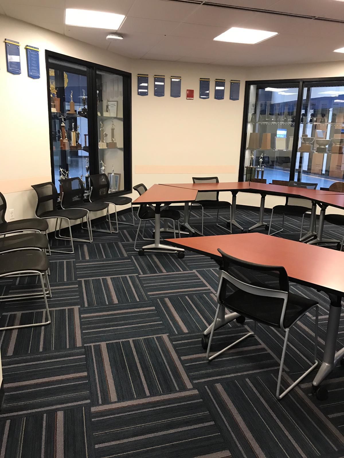 A classroom with a u-shaped table, chairs, and awards behind glass doors. Dark blue and gray carpet.