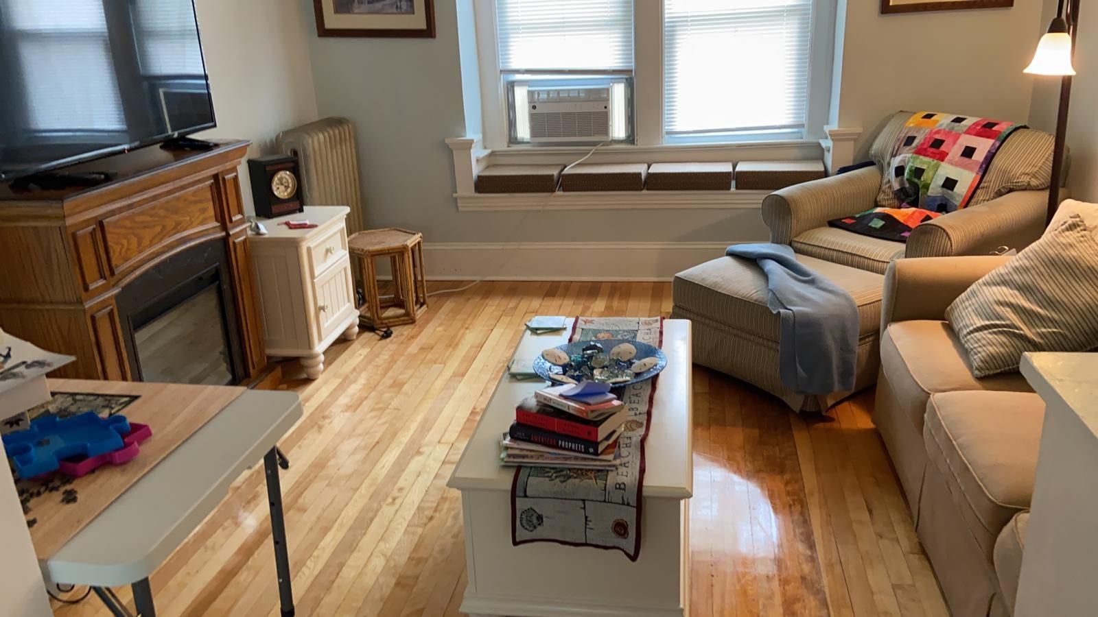 Cozy living room with fireplace, couch, armchair, and window seat. Hardwood floors and natural light.
