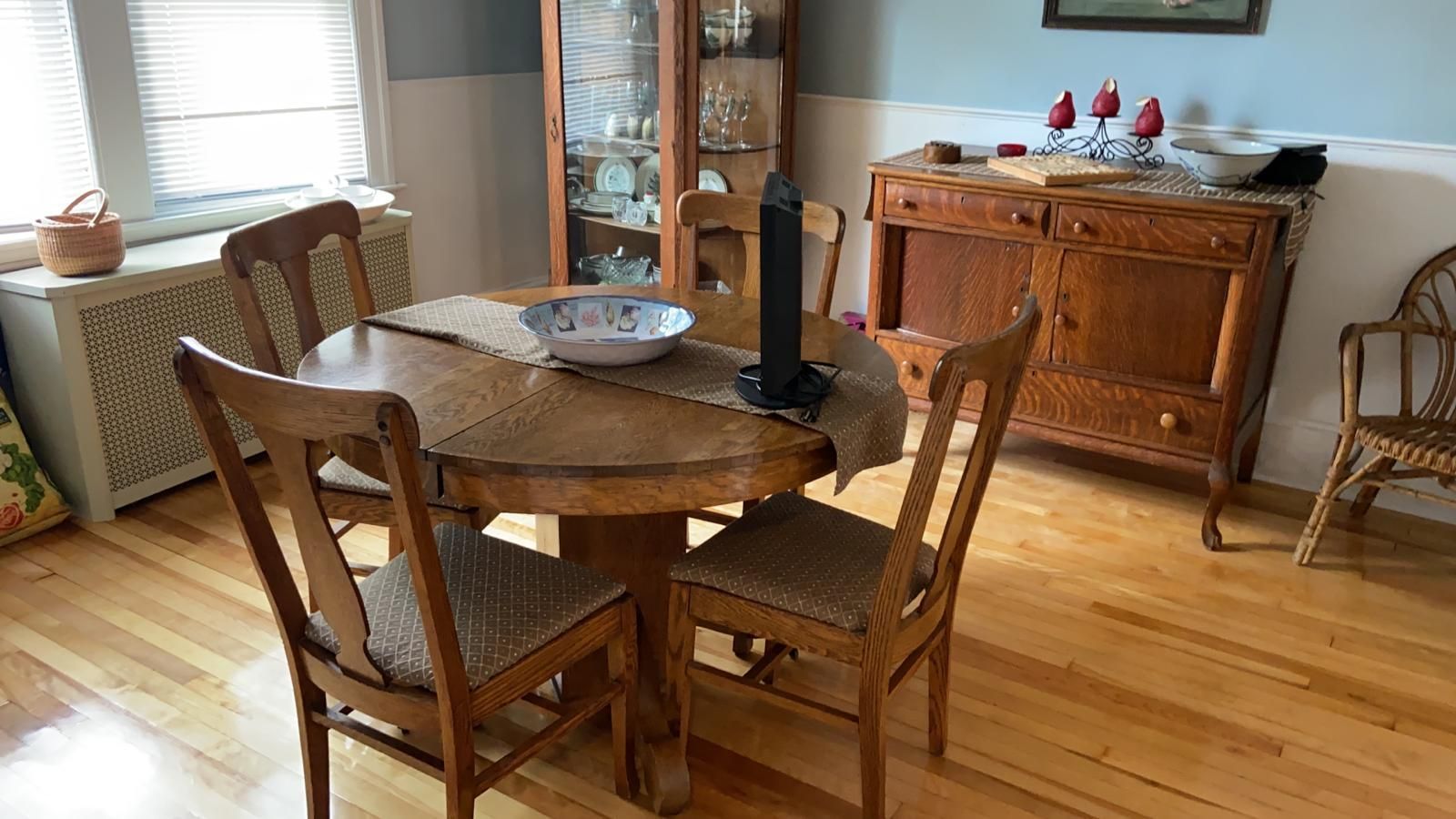 A dining room with a wooden table, chairs, and buffet. Sunlight streams through a window.